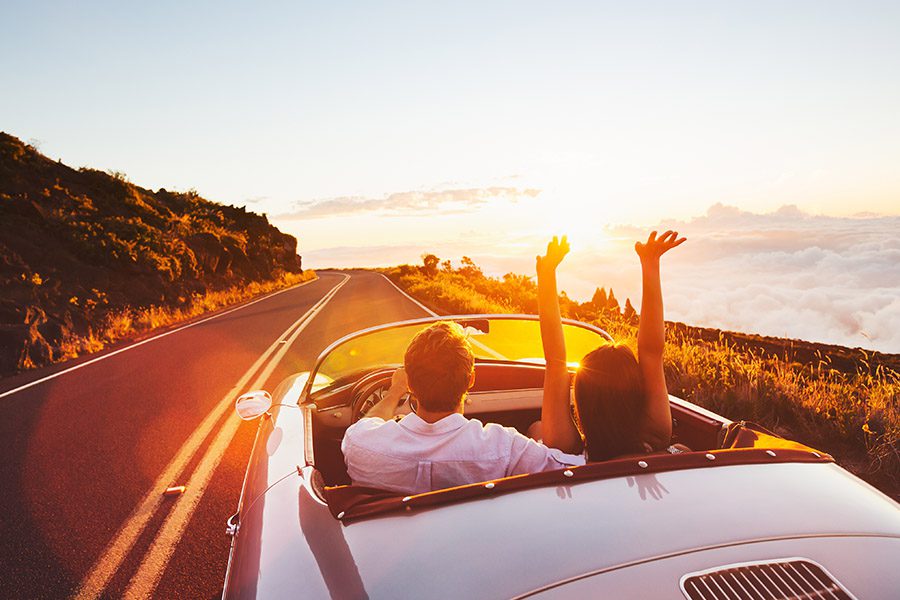 Personal Insurance - Couple Driving Down a Road in a Vintage Convertible Car and Female Rider Has Her Arms Raised at Sunset